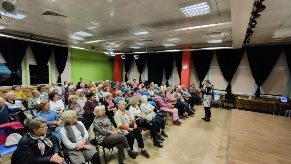zdjęcie przedstawia grupę siedzących seniorów i kobietę prowadzącą wykład