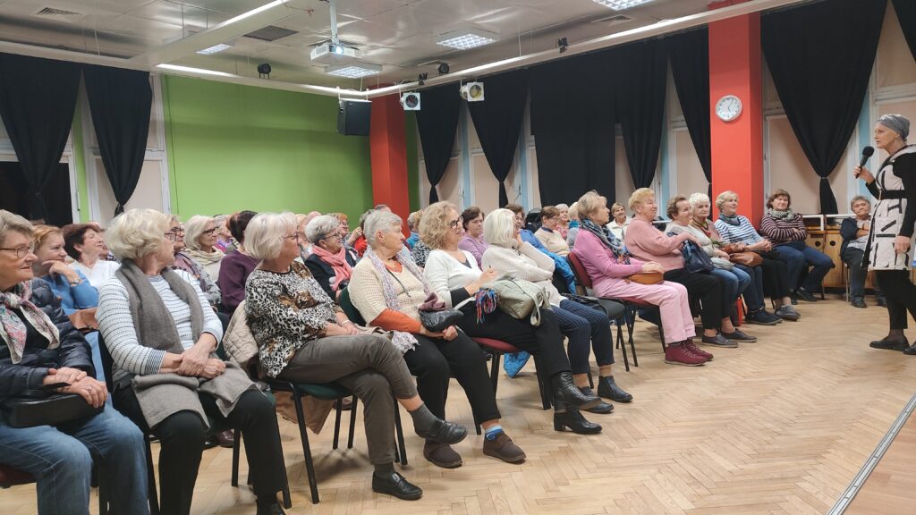 zdjęcie przedstawia grupę siedzących seniorów i kobietę prowadzącą wykład