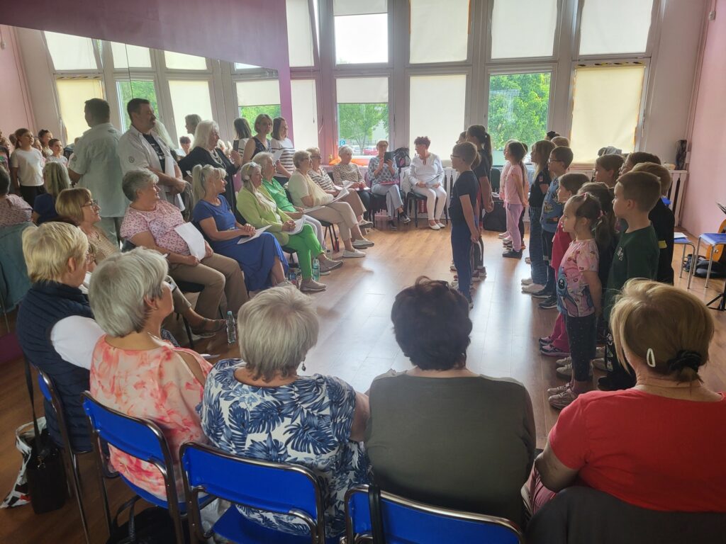 Zdjęcie przedstawia dzieci i seniorów biorących udział w próbie przed koncertem z okazji Dnia Matki w klubie "Dąb"