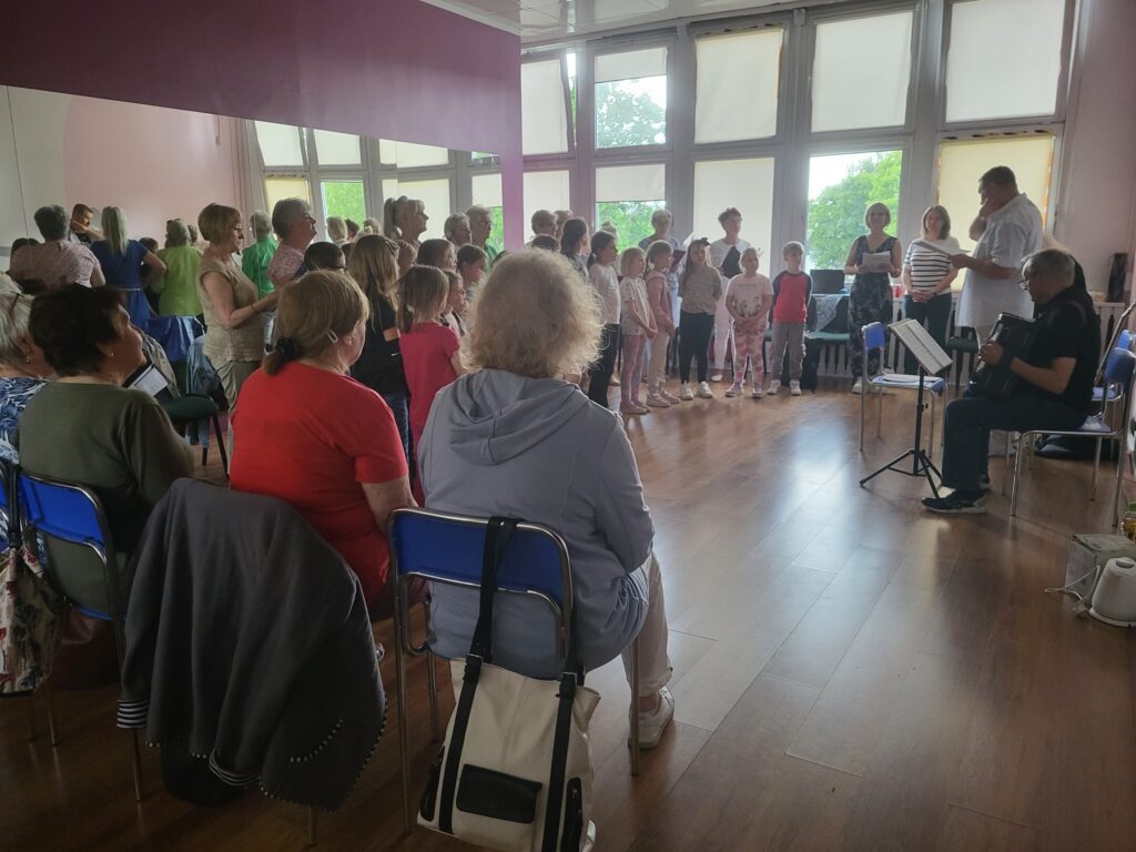 Zdjęcie przedstawia dzieci i seniorów biorących udział w próbie przed koncertem z okazji Dnia Matki w klubie "Dąb"